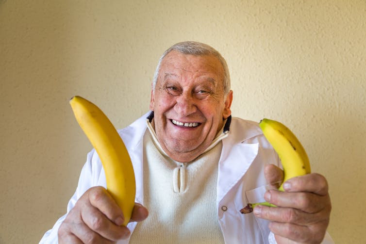 Older smiling man holding banana in each hand, one large, one small