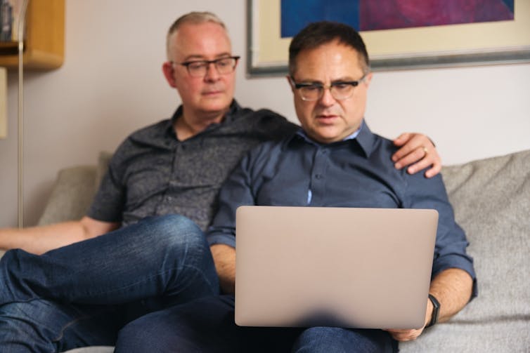 Older gay couple sitting on sofa, one with hand on shoulder, looking at open laptop