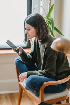 A young woman sits on a chair, looking at a phone.