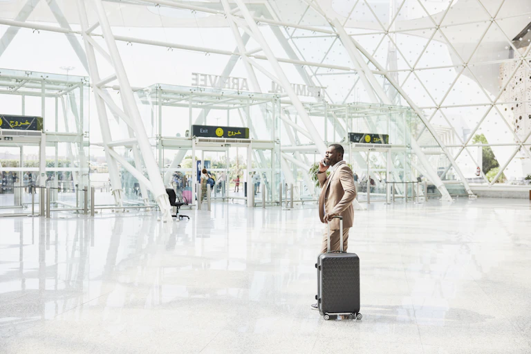 Wide shot of a man wearing a suit, standing with a suitcase and using a phone in an airport terminal.