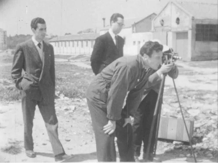 Cuatro jóvenes están en un descampado, mirando fuera del encuadre, mientras uno observa algo por una cámara de cine.