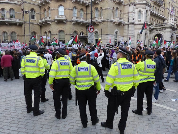 Five met police officers in hi-vis jackets stand near a crowd of pro-Palestine marchers.
