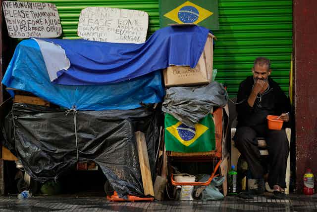 Homem de meia-idade alimenta-se de uma vasilha ao lado de caixas, sacos plásticos e bandeiras do Brasil numa calçada de grande cidade