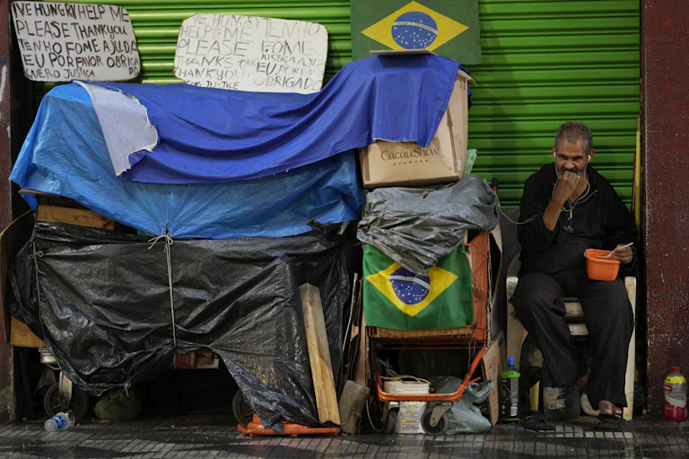 Homem de meia-idade alimenta-se de uma vasilha ao lado de caixas, sacos plásticos e bandeiras do Brasil numa calçada de grande cidade