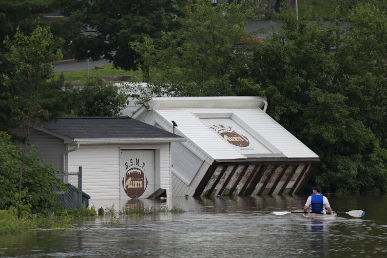 A small white building lies tilted on its back in floodwater as someone kayaks past it.
