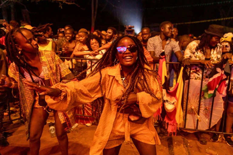 A crowded audience dancing at night, in the foreground a smiling young woman in shades.