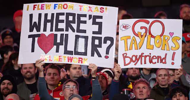 Football fans hold up signs that read "I flew from Texas! Where's Taylor?" and "Go Taylor's Boyfriend!" in the stands at a Kansas City Chiefs game.