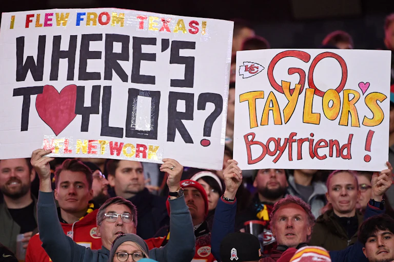 Football fans hold up signs that read "I flew from Texas! Where's Taylor?" and "Go Taylor's Boyfriend!" in the stands at a Kansas City Chiefs game.