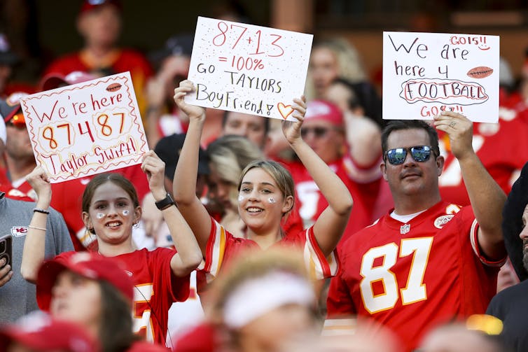Three fans hold signs at an NFL game. One, a young girl's, says, 