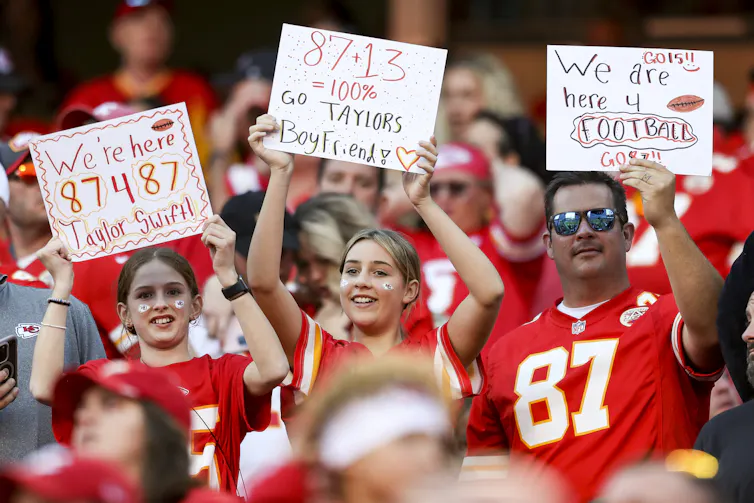 Three fans hold signs at an NFL game. One, a young girl's, says,