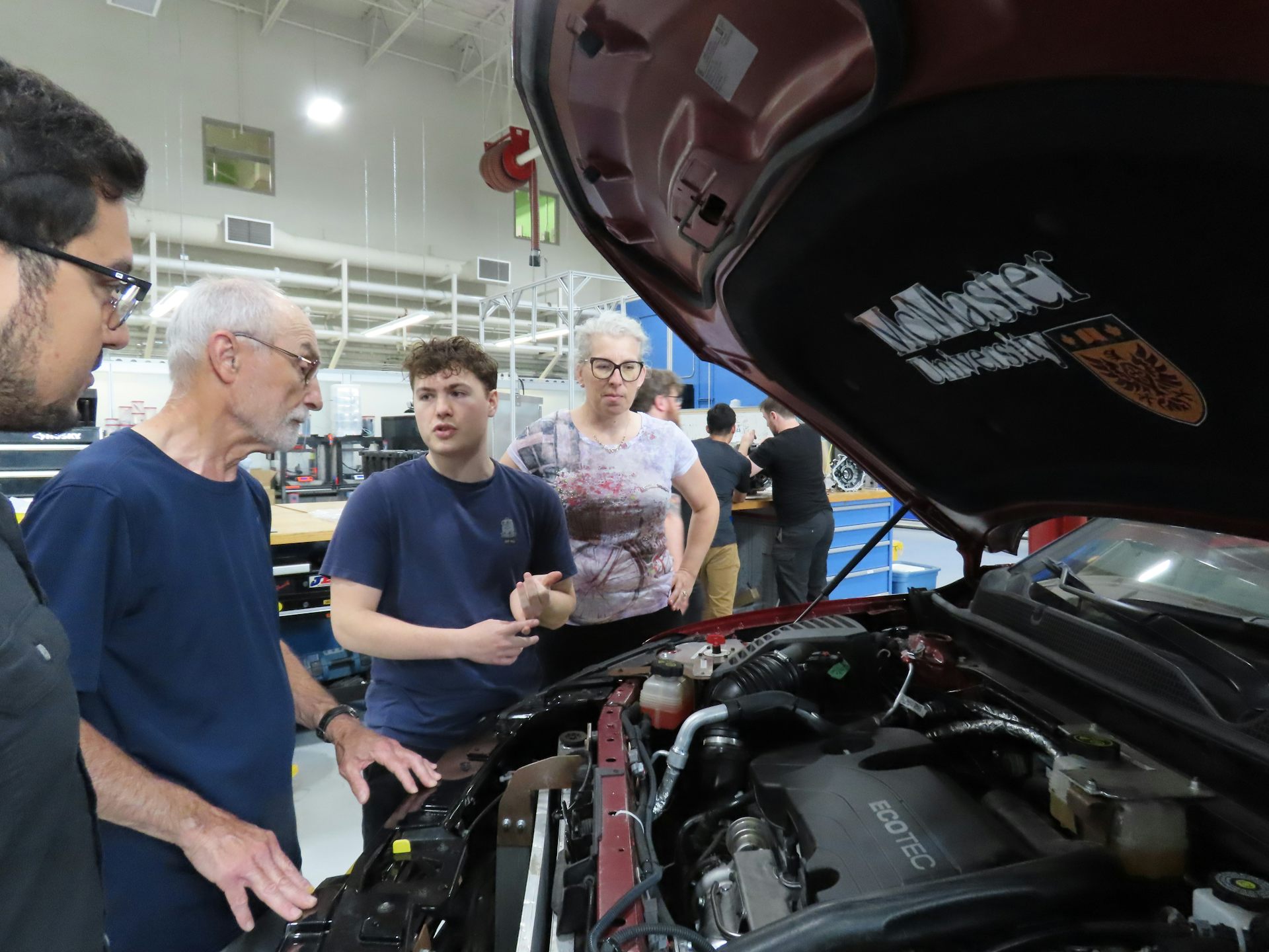 An older adult, researcher Brenda Vrjklan and two students look under the hood of a car in a lab or workshop