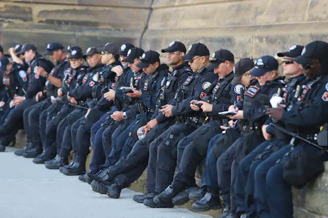 Uniformed police officers sit in a row, most looking at their phones.