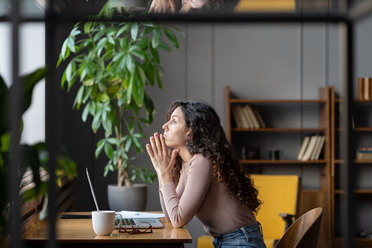 A woman sitting at a desk and looking out a window with a serious expression on her face.