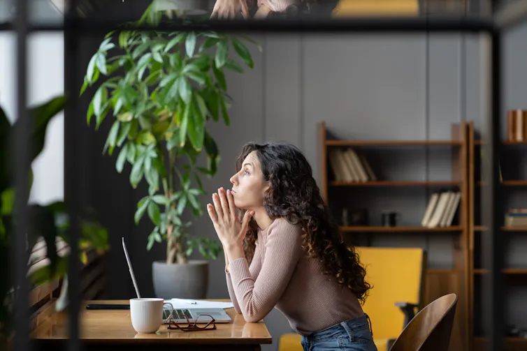 A woman sitting at a desk and looking out a window with a serious expression on her face.