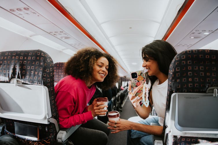 Two passengers chat and look at a phone screen on a plane.
