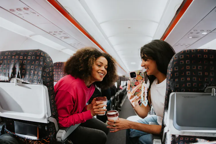 Two passengers chat and look at a phone screen on a plane.
