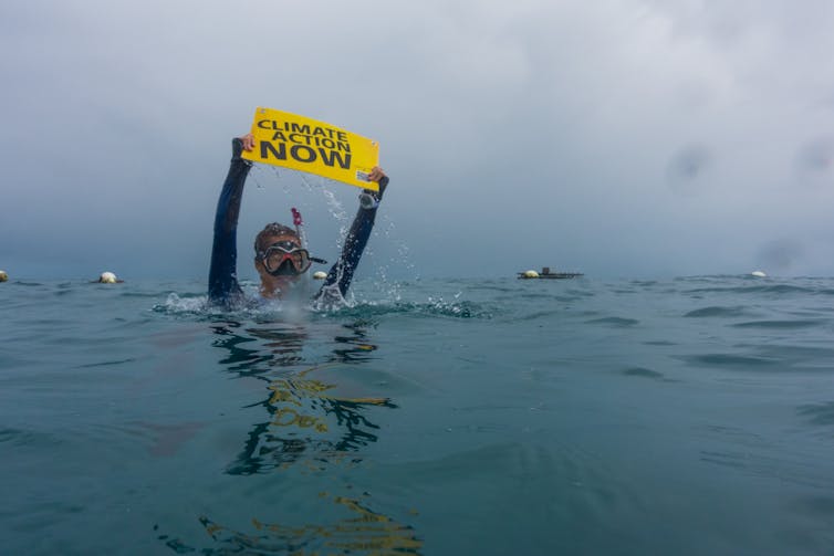 A partially submerged diver on the Great Barrier Reef, holding aloft a yellow sign bearing the words 'Climate Action NOW'