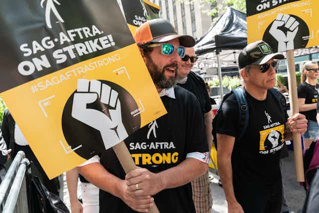 Dos hombres con camisetas negras, gorras y gafas de sol se manifiestan, portando pancartas sobre la huelga de actores en Hollywood, junto a otros compañeros.