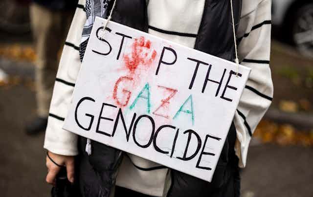 Uma pessoa, vista apenas do pescoço para baixo, segura um cartaz que diz "Stop the Gaza Genocide" (Pare o genocídio de Gaza), com uma mão vermelha substituindo o "o" da palavra stop.