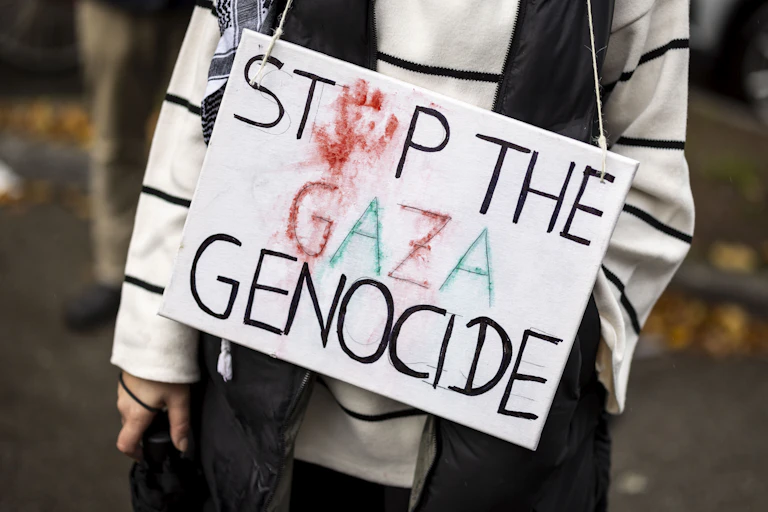 Uma pessoa, vista apenas do pescoço para baixo, segura um cartaz que diz "Stop the Gaza Genocide" (Pare o genocídio de Gaza), com uma mão vermelha substituindo o "o" da palavra stop.
