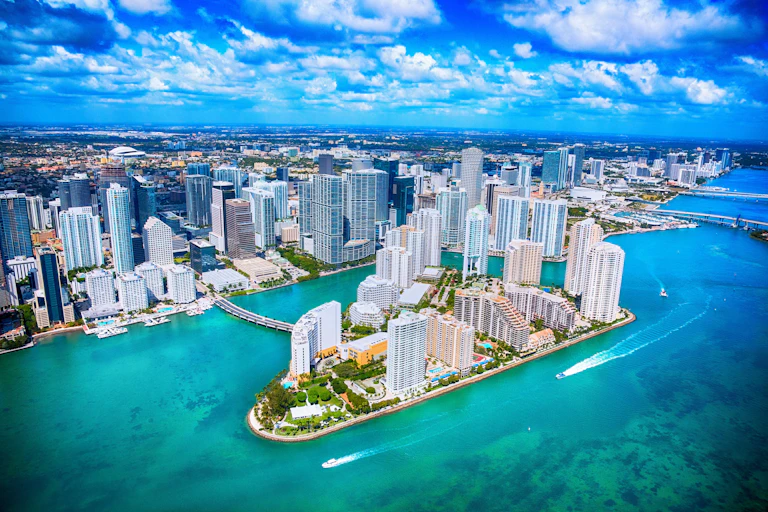 A helicopter of view of Miami's gleaming office towers over Biscayne Bay, where boats are running through the channels.