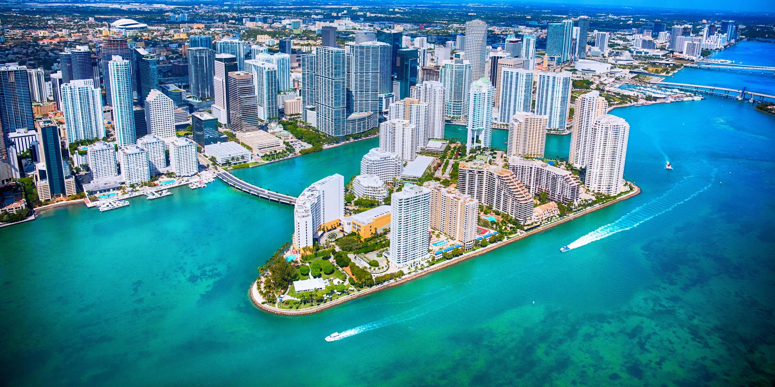 A helicopter of view of Miami's gleaming office towers over Biscayne Bay, where boats are running through the channels.