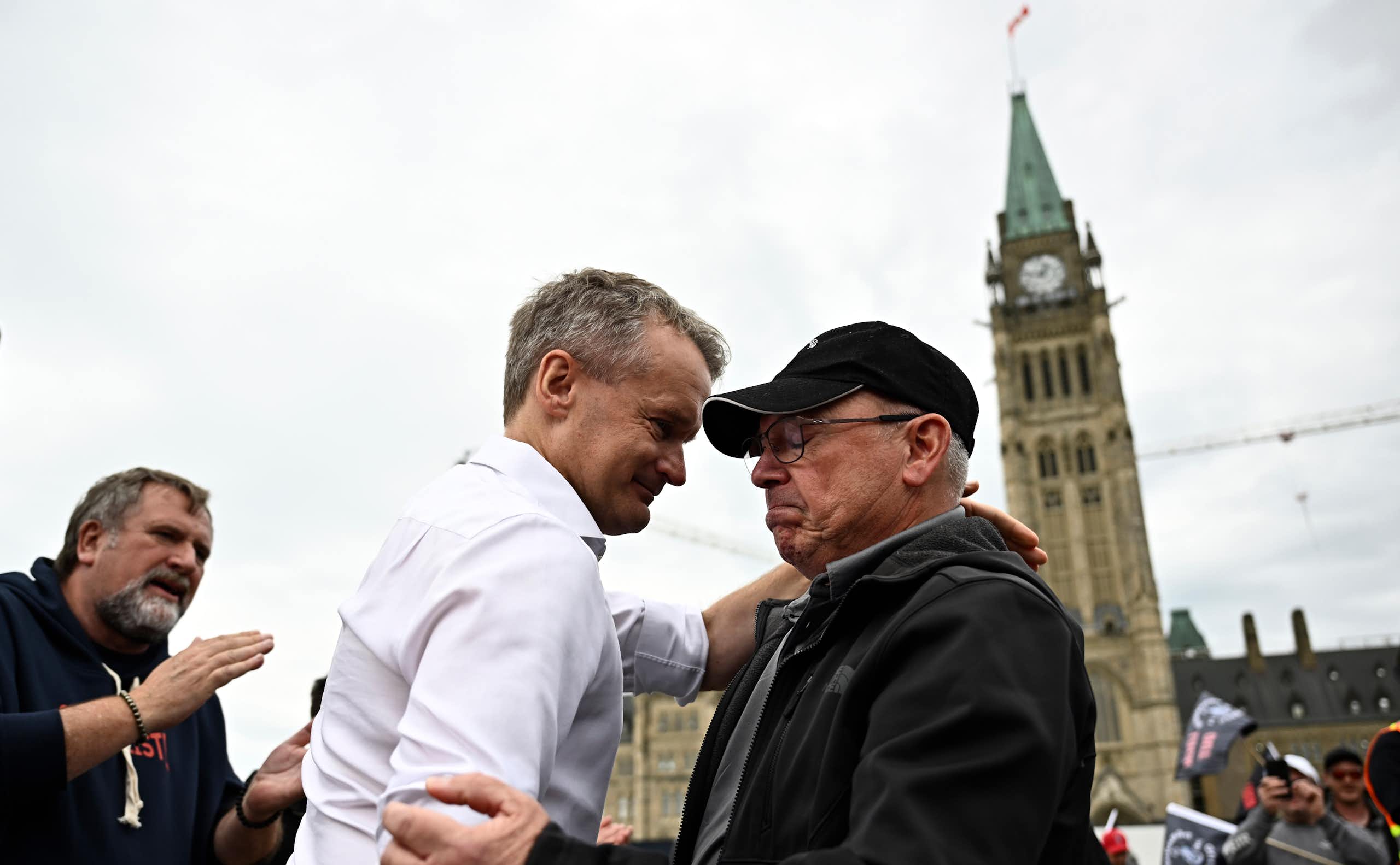 A man in a white shirt and tie embraces an older man in a ball cap with the Peace Tower behind them.