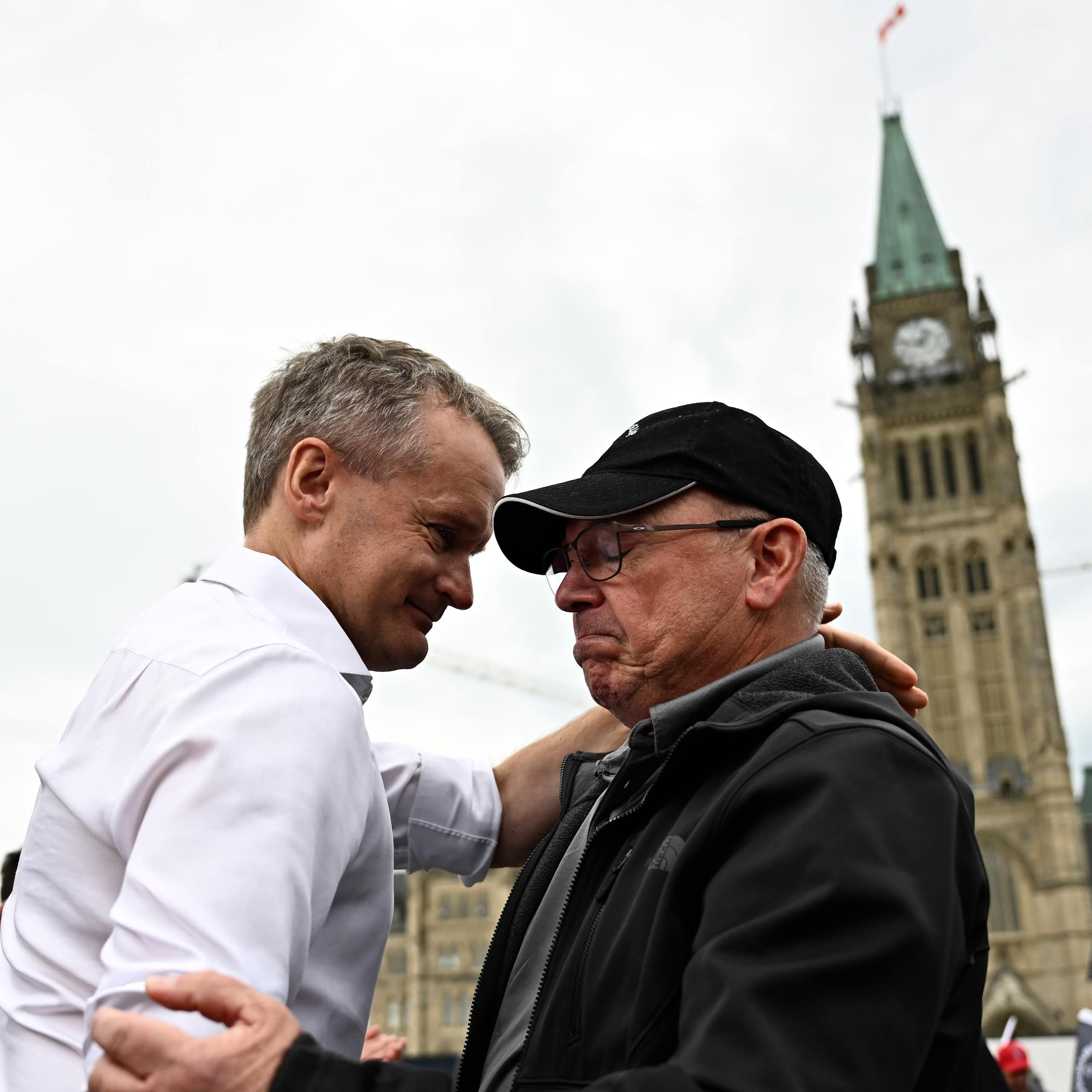 A man in a white shirt and tie embraces an older man in a ball cap with the Peace Tower behind them.