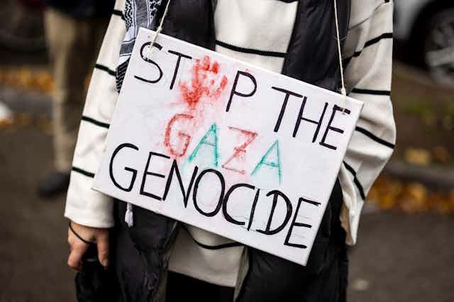 A person, only seen from the neck down, holds a sign that says 'Stop the Gaza Genocide' with a red hand print standing in for the o in the word stop.