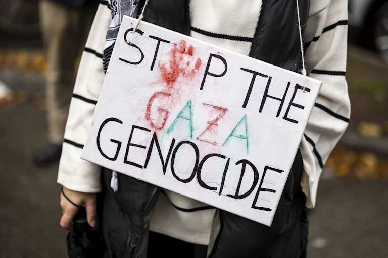 A person, only seen from the neck down, holds a sign that says 'Stop the Gaza Genocide' with a red hand print standing in for the o in the word stop.