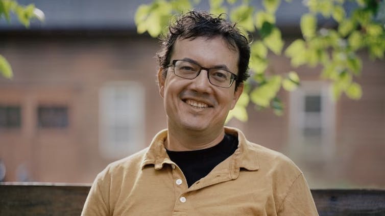 A man with glasses and dark hair, smiling, wearing a polo shirt over a black t-shirt. Leafy branches in background.