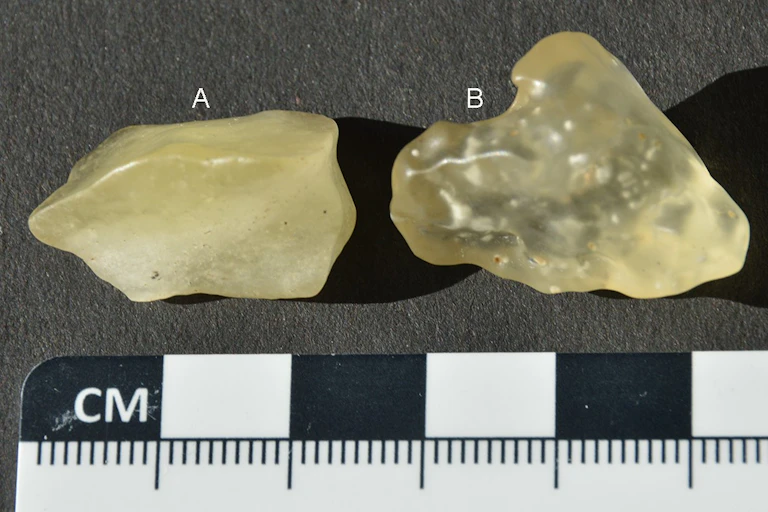 A photograph of two yellow stones - one appears almost translucent and the other is a deeper yellow - against a black background, with a small ruler beneath them for scale