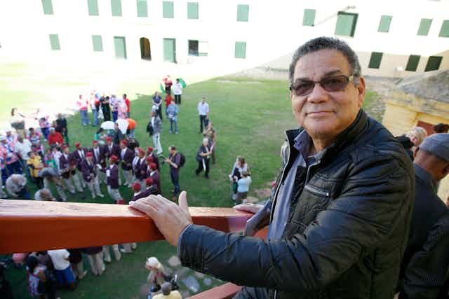 A man stands on a staircase looking into camera with a slight smile. He wears sunglasses and a black leather jacket. On the grass below, a crowd of people gathers, some dressed in Cape minstrel outfits.