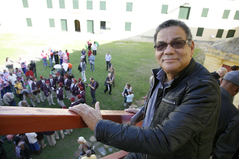 A man stands on a staircase looking into camera with a slight smile. He wears sunglasses and a black leather jacket. On the grass below, a crowd of people gathers, some dressed in Cape minstrel outfits.