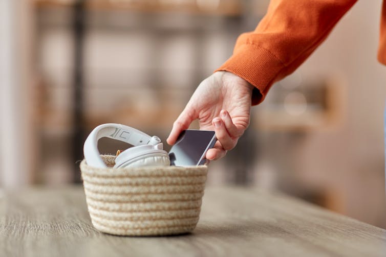 A person puts their smartphone into a basket next to their headphones.