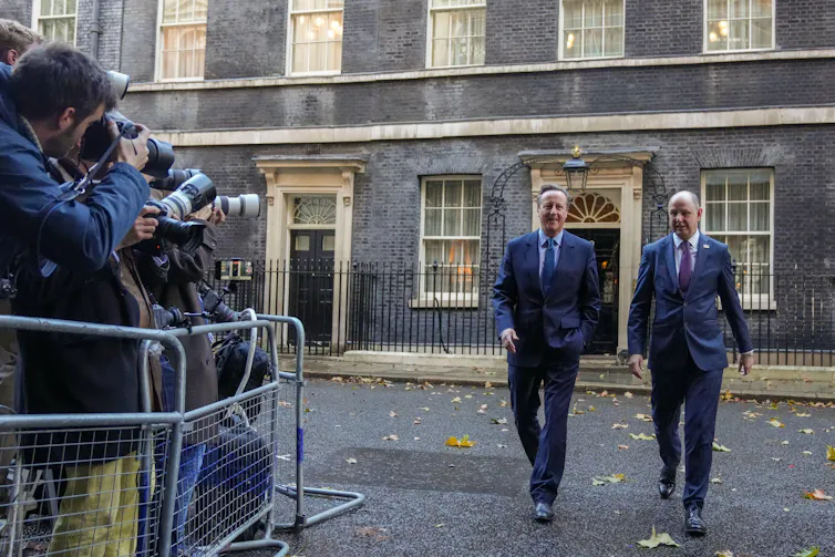 David Cameron and another man walk past a group of news photographers.