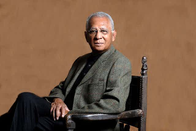 A man sits for a formal portrait in a blazer, greying hair and glasses on. The chair has African carvings on it.