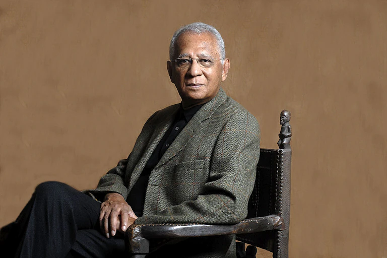 A man sits for a formal portrait in a blazer, greying hair and glasses on. The chair has African carvings on it.