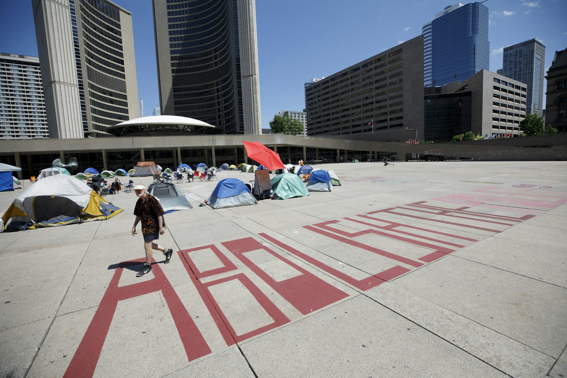 A man walks past red painted letters on the pavement reading Abolish the Police outside Toronto's City Hall on a sunny day.