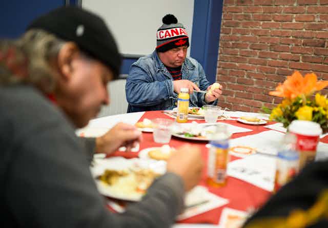 A man in a Canada toque butters a roll. Another man is seen out of focus eating in the foreground.
