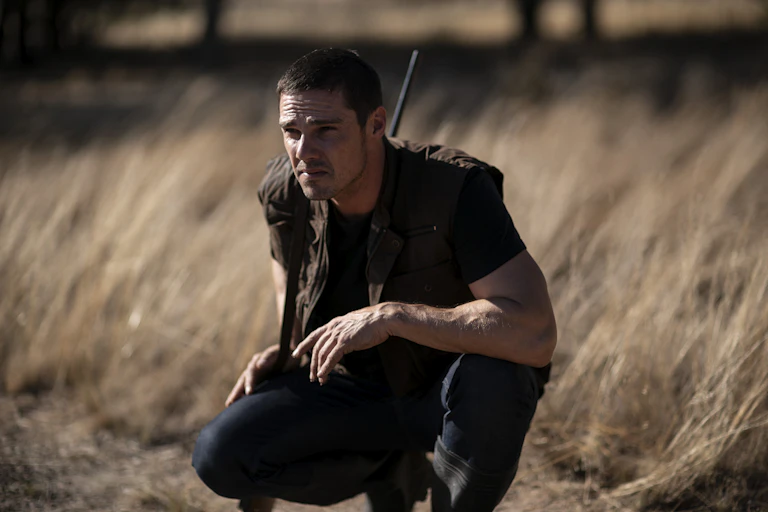 A man with a gun crouches among dead grasses