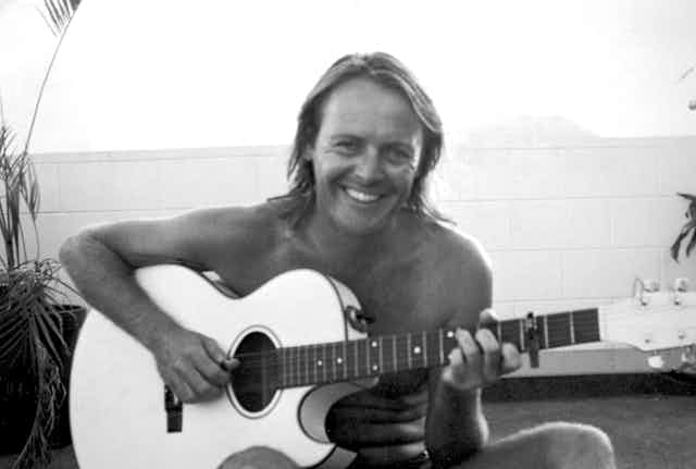 A shirtless man holding a guitar smiles at the camera