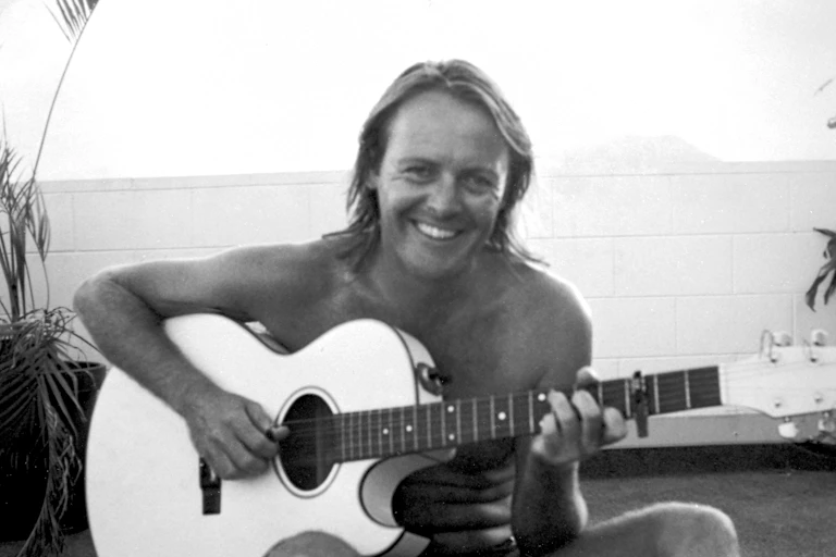 A shirtless man holding a guitar smiles at the camera