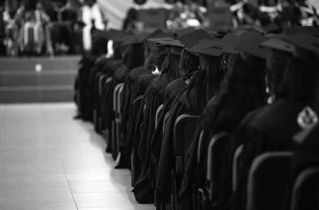 Students in academic dress sit in rows, at a graduation ceremony. 