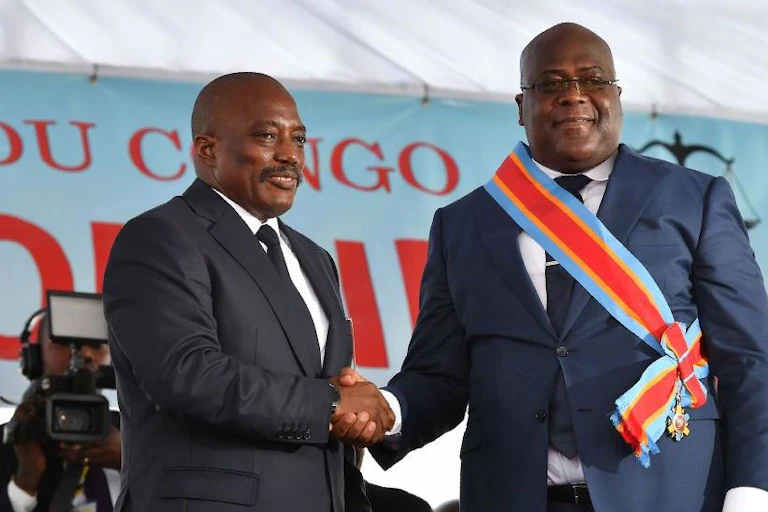 Two men shaking hands with a video camera behind them.