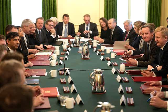 Cabinet ministers seated around a long table, each with a red folder and name placard in front of them. Rishi Sunak and David Cameron are seated opposite each other.