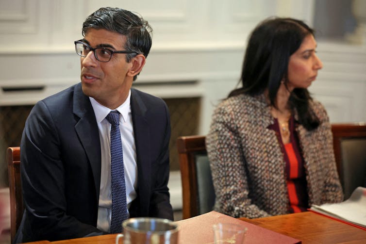 Rishi Sunak and Suella Braverman sitting side by side at a table, looking in opposite directions. Sunak is in focus while Braverman is out of focus.