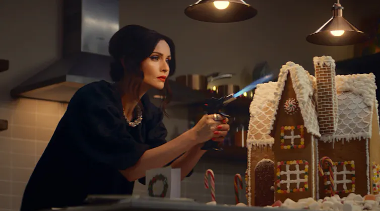 A woman holds a blow torch in a kitchen, in front of a gingerbread house.