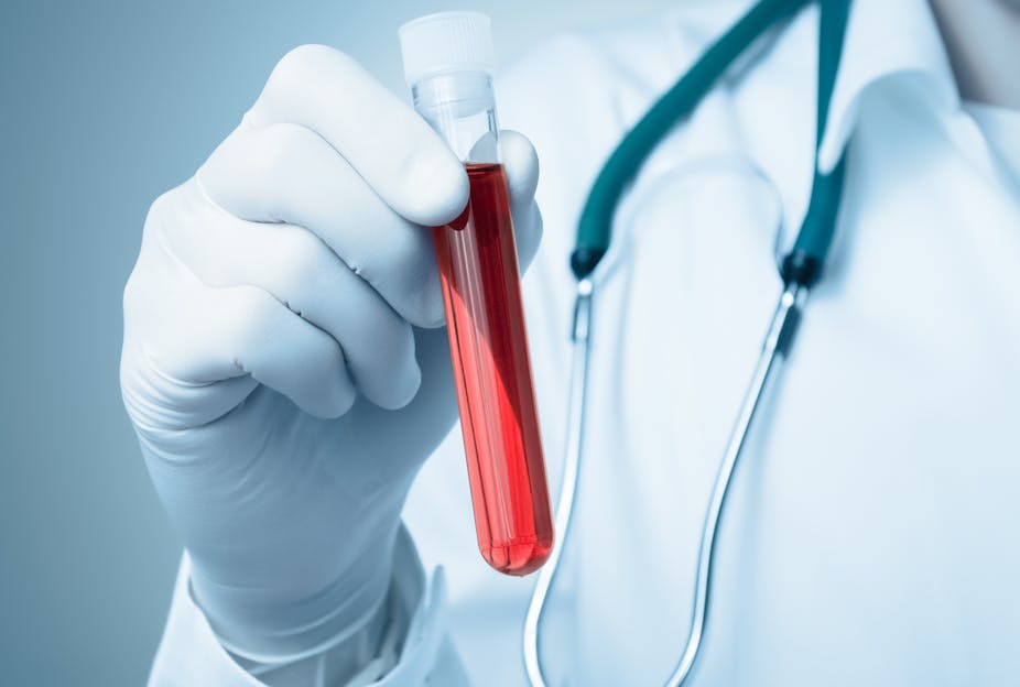 La main d'un professionnel de santé gantée tient un tube à essai contenant un liquide rouge évoquant le sang.