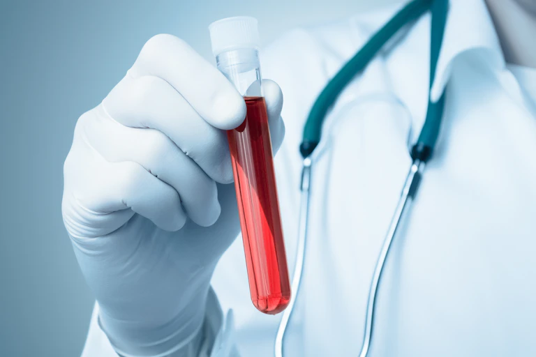 La main d'un professionnel de santé gantée tient un tube à essai contenant un liquide rouge évoquant le sang.